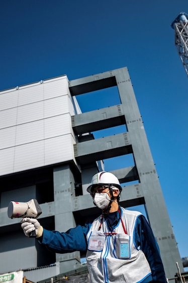 a person in a mask and a hard hat holds a geiger counter to measure radiation in front of the Fukushima nuclear power plant