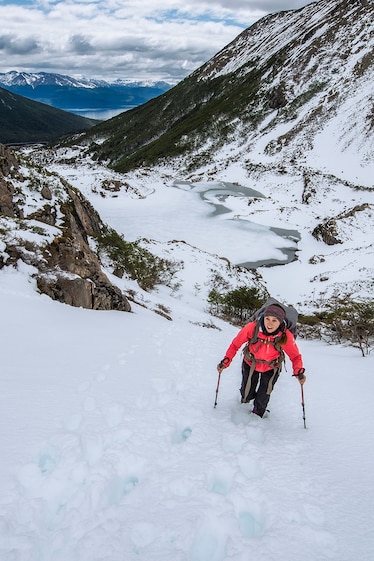 A hiker outfitted with winter gear, hiking poles, and a full pack, ascends a snowy incline. Mountaintops fill the horizon behind her.