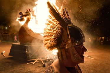 a man in a viking feathered helmet in front of a burning longship